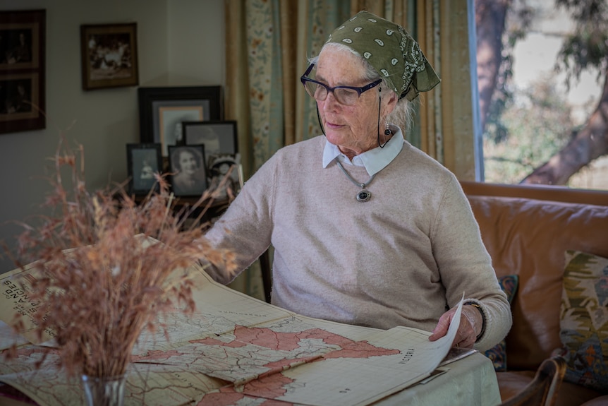 A woman sits at a table reading a map, wearing a beige jumper and green bandana.