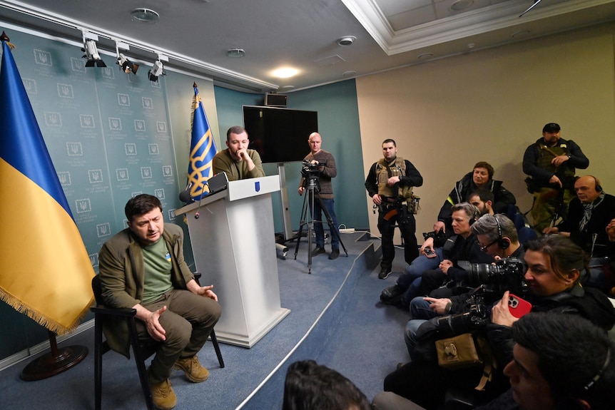 Zelenskyy sits on a podium surrounded by media and men holding guns. A Ukrainian flag is behind him. 