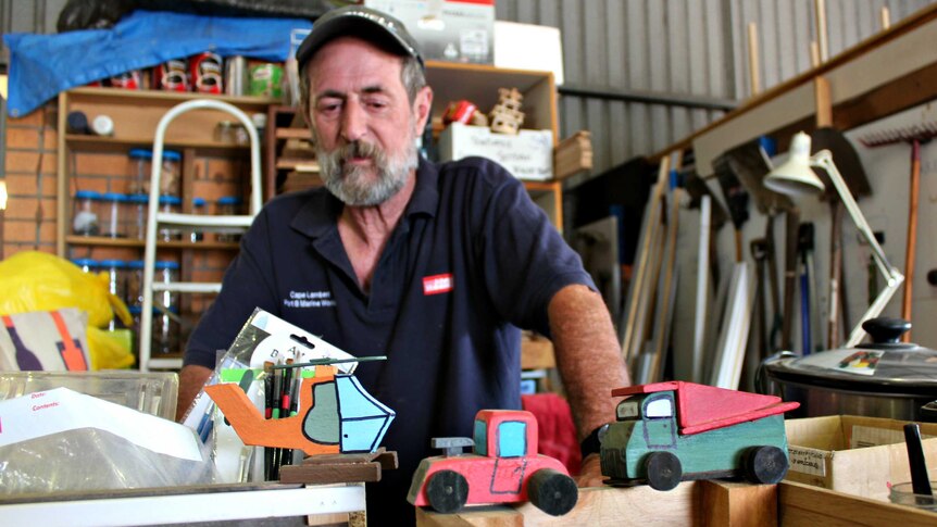 Dave Smith standing in Men's Shed workshop behind colourful handmade wooden helicopter and trucks.