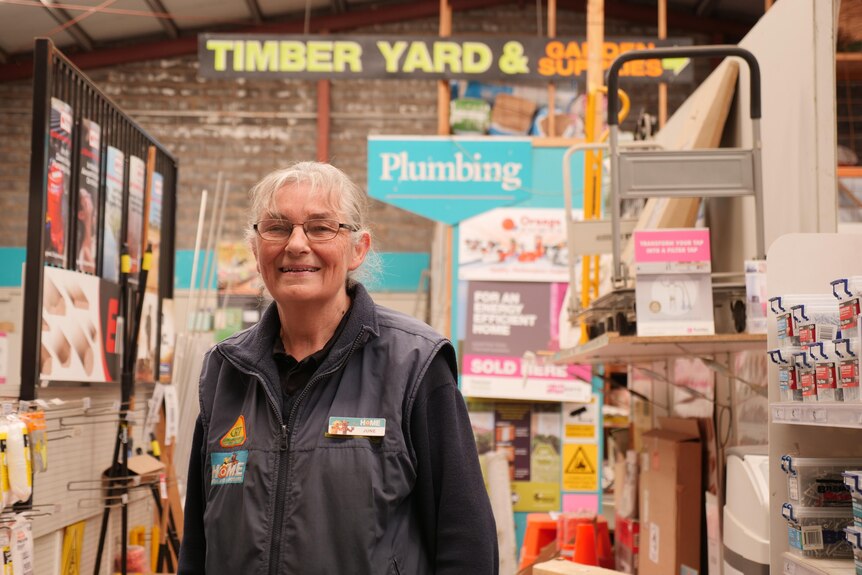 A woman of retirement age stands in front of hardware warehouse with sign for timber yard.