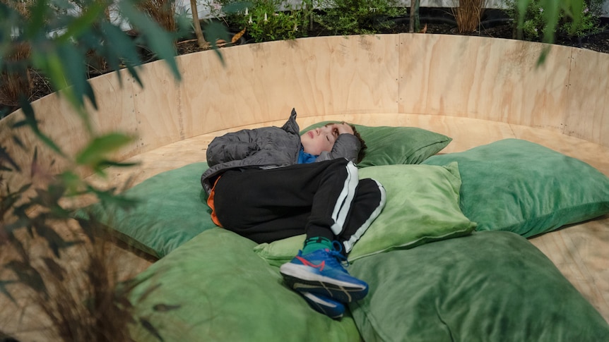 A child lies down, eyes closed, in a round wooden space covered in green cushions, surrounded by green leafy plants. 