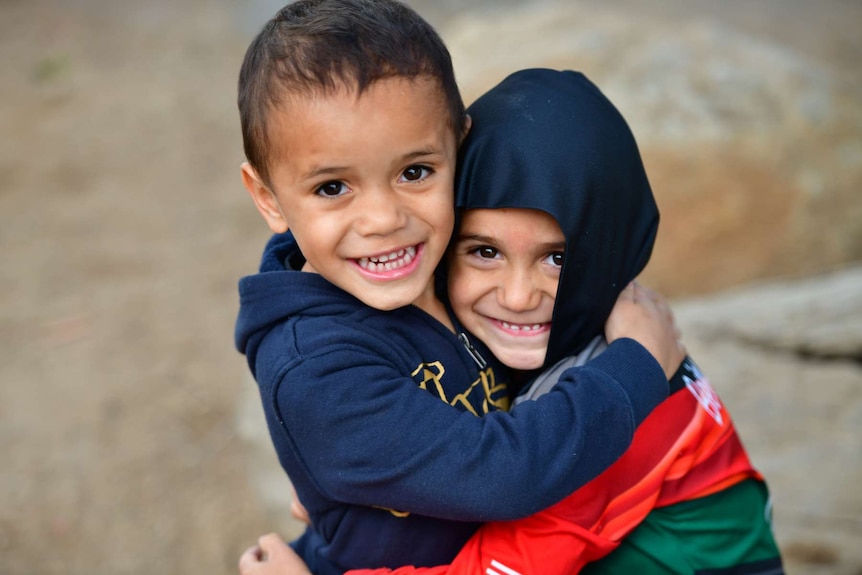 Two young Indigenous boys wearing jumpers hugging and smiling as they look directly into the camera.