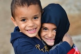 Two young Indigenous boys wearing jumpers hugging and smiling as they look directly into the camera.