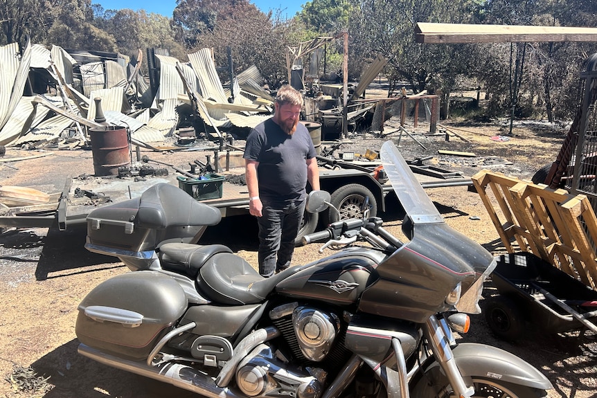 Damon Hendricksen looks at a vintage motorcycle that sits in front of the ruins of a home on a burnt property.