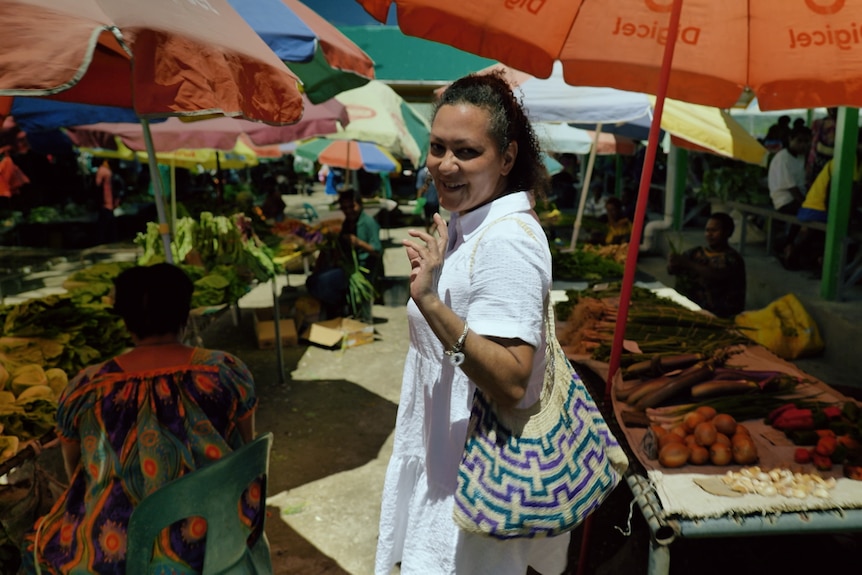 Woman wearing white dress standing in the middle of an outdoor fruit and vegetable market. 