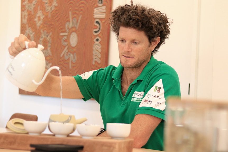 A man with a mop of brown curly hair pours a pot of tea into a cup while wearing a green polo shirt.