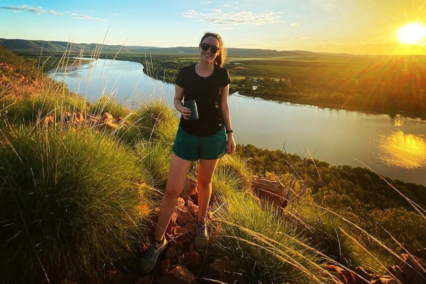 A woman wearing a t-shirt, shorts and sunglasses smiles on a river bank as the sun sets.