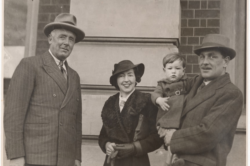 Black and white photo of two men in suits and hats, one holding a small child and a woman in coat and hat.