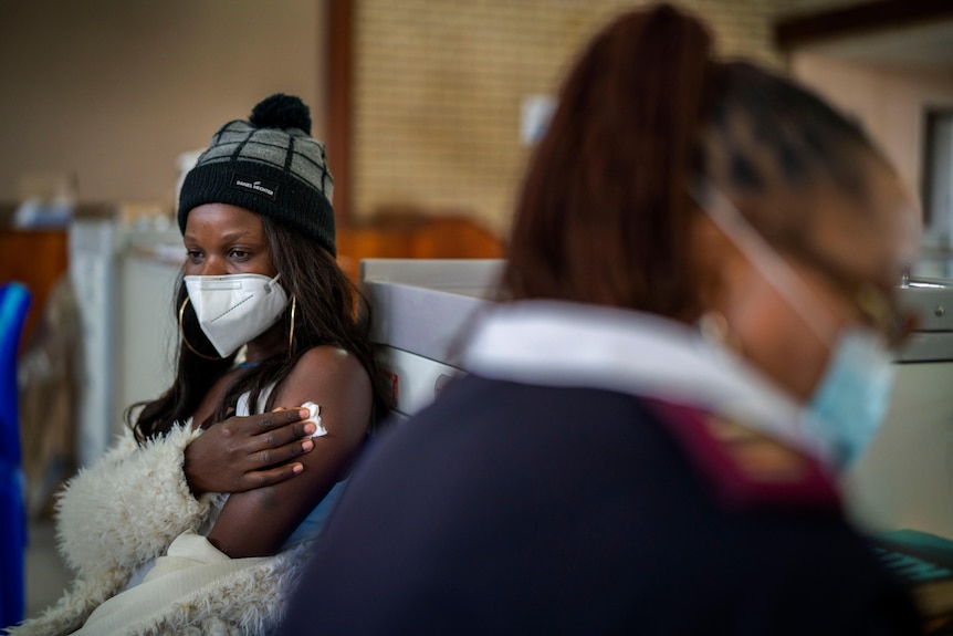 A woman in a beanie and face mask holds a cotton ball to her shoulder, looking deep in thought