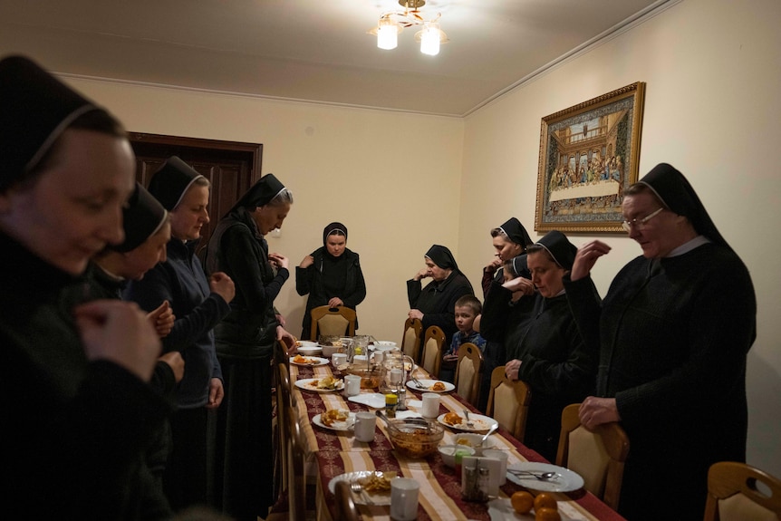 A group of nuns stand around a table adorned with food, making signs of the cross with their hands.
