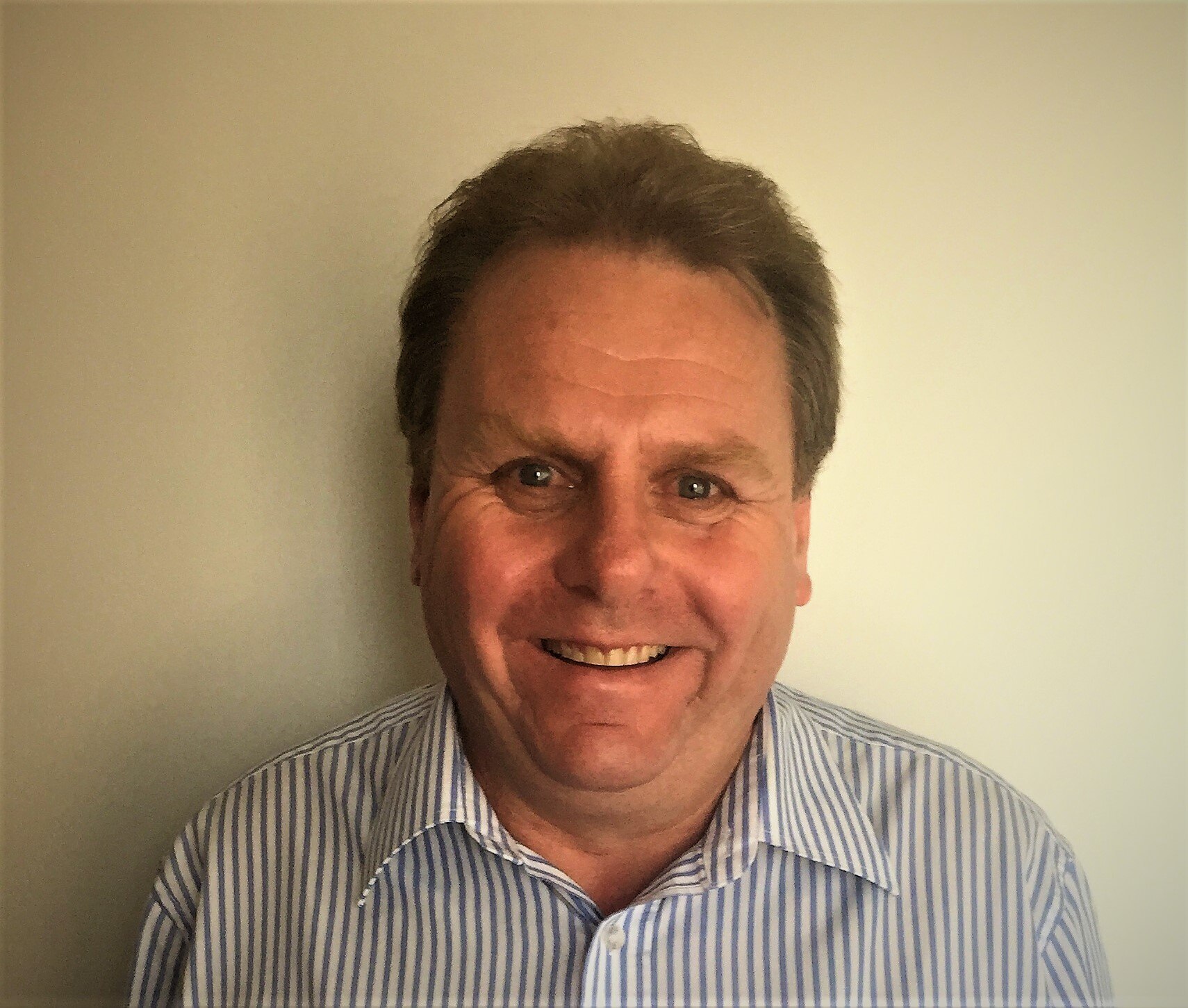 A dark-haired man wearing alight-coloured shirt stands against a wall, smiling.