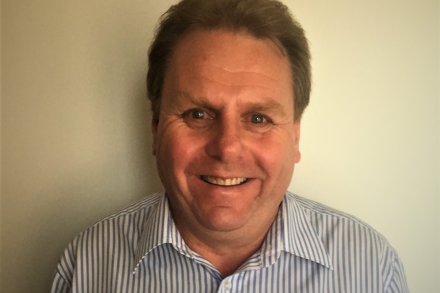 A dark-haired man wearing alight-coloured shirt stands against a wall, smiling.