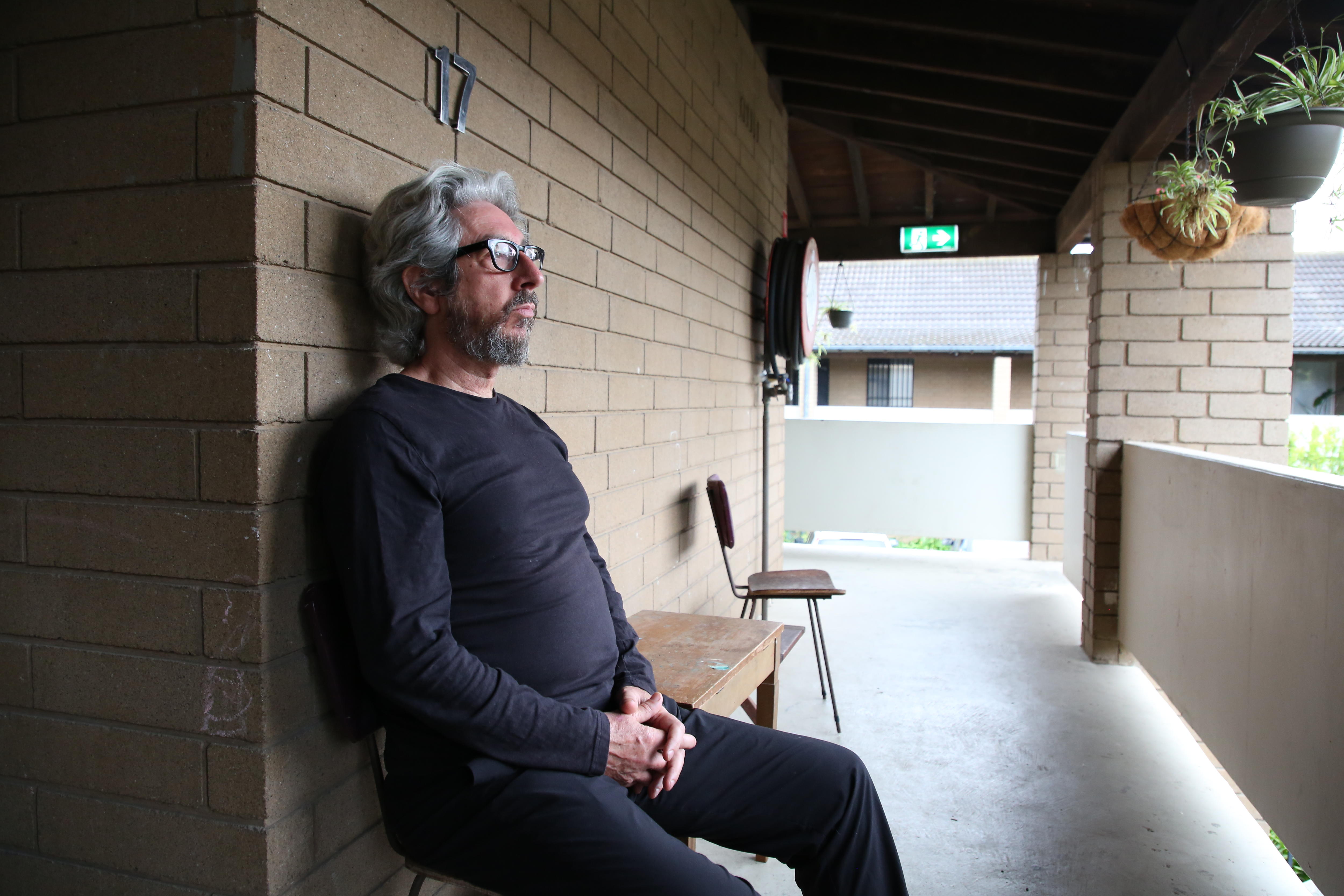 John sitting on his porch with his back against a brick wall, looking very stoic.