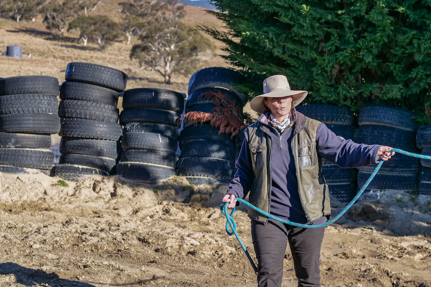 A woman wearing a broad-rimmed hat walks and spins a rope inside a horse pen.
