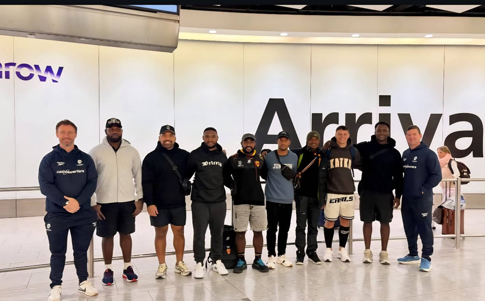 A group of smiling men in hoodies stand together at an airport.