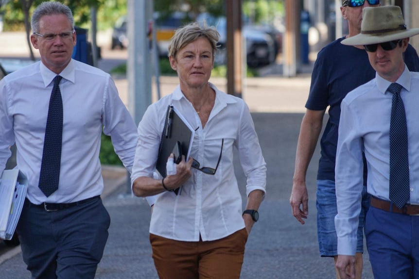 A woman flanked by two men heading to court walking outside on a Darwin street with a white shirt.