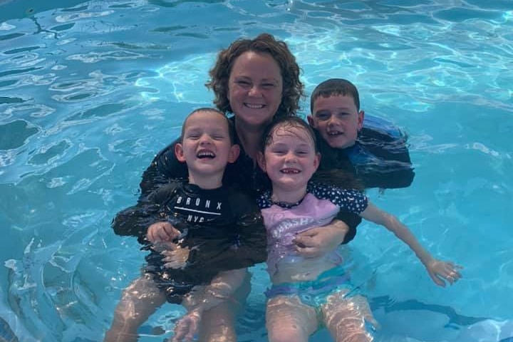 A mother with her arms around her three young children in a swimming pool