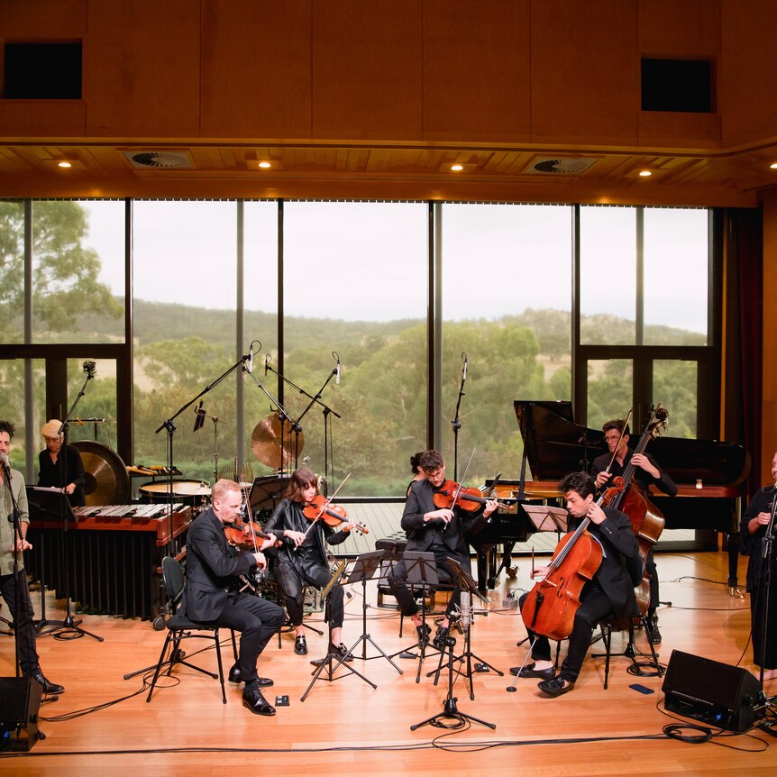 Lior and Dr Bennett on stage with musicians a large window in the background show Adelaide Hills bush.