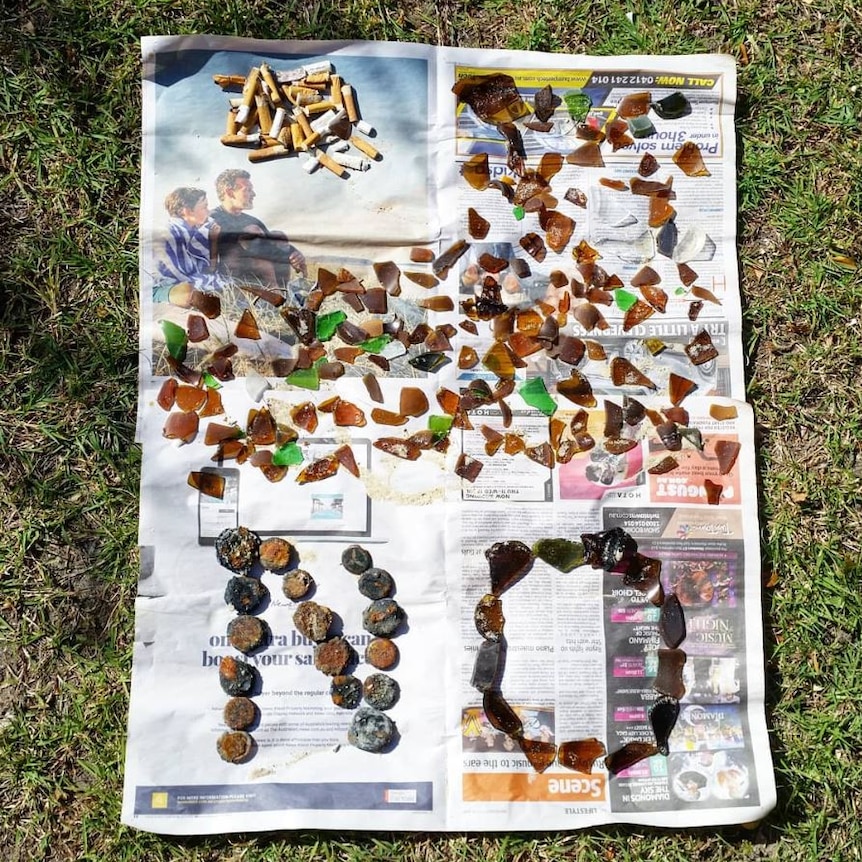 Broken pieces of brown and green glass, cigarette butt are placed on newspaper while rusty beer bottle lids form the word 'No'