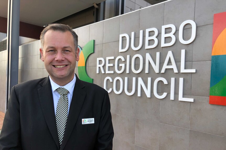 A middle-aged man in a dark suit smiling as he stands in front of a sign reading "Dubbo regional Council".