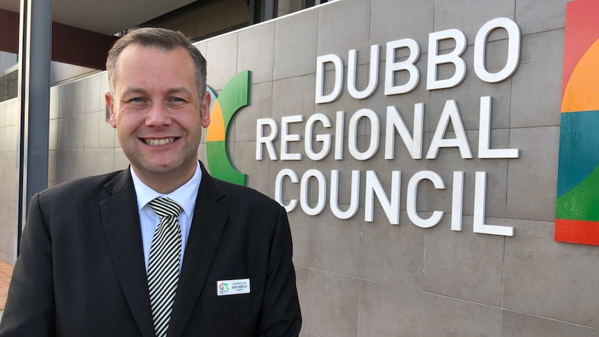 A smiling man in a dark suit stands in front of a wall bearing the lettering "Dubbo Regional Council".