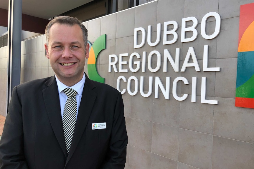 A smiling man in a dark suit stands in front of a wall bearing the lettering "Dubbo Regional Council".