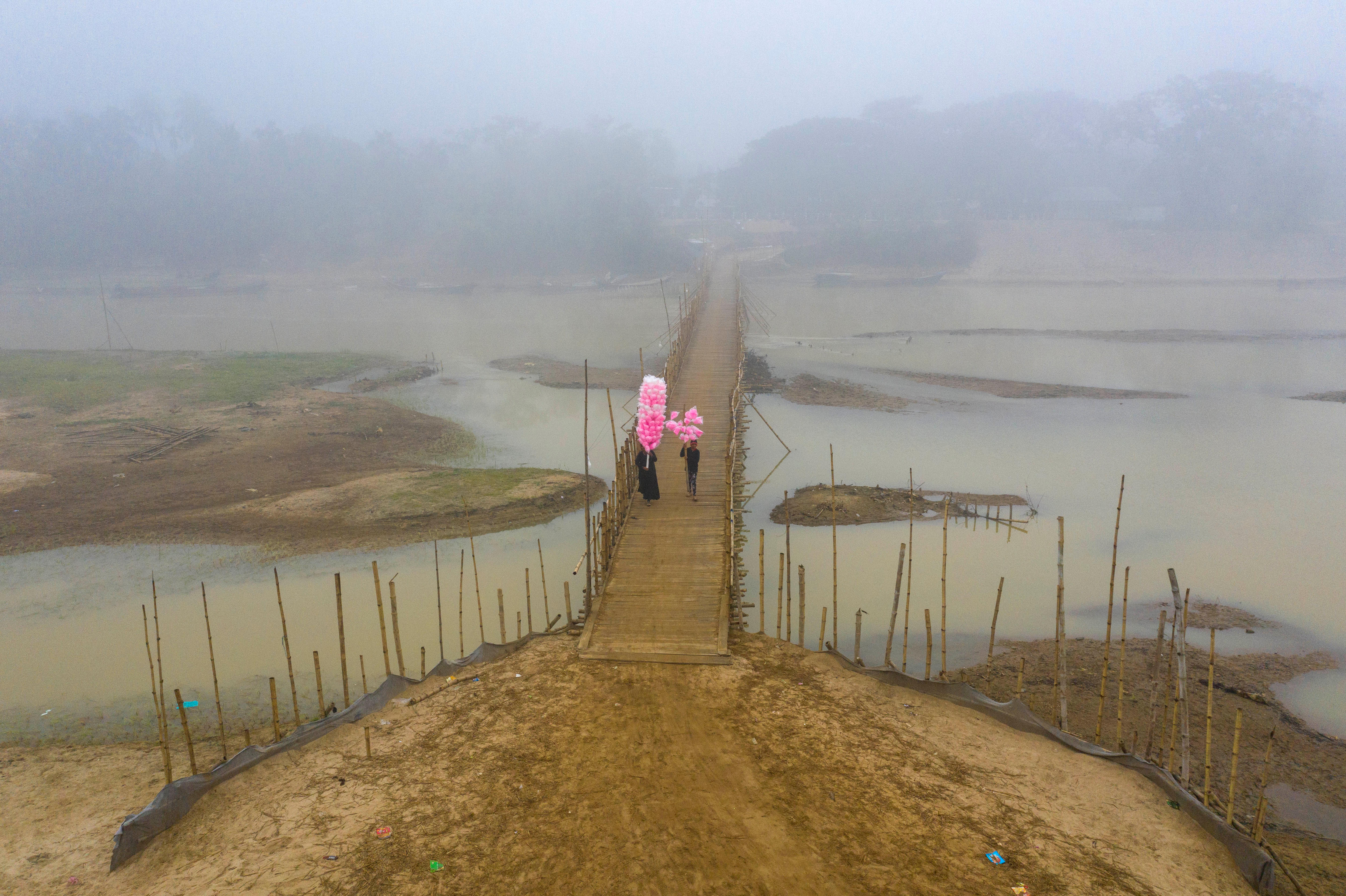 A woman and child carry bright pink sweets, which stand out against cloudy water and fog, tied to poles across a bridge. 
