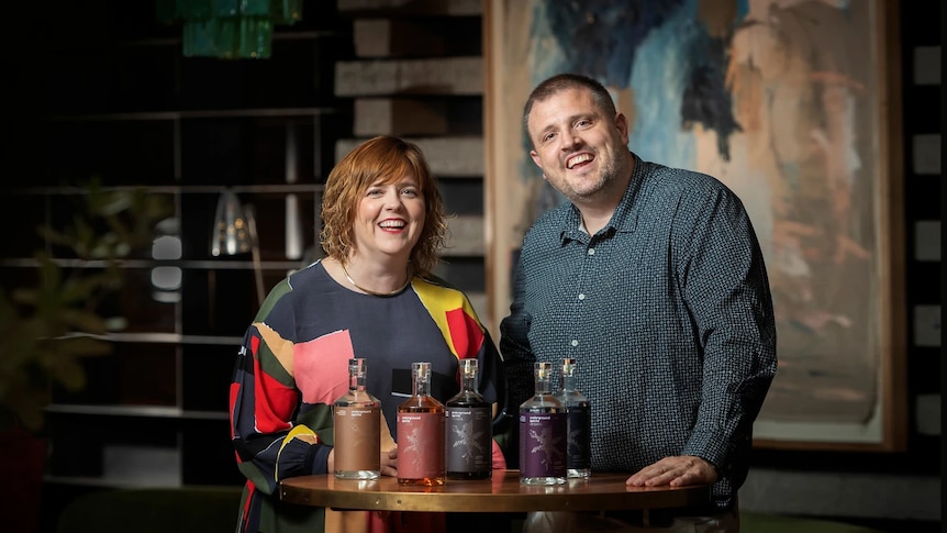 A woman and a man smile in front of a table with several gin and vodka bottles