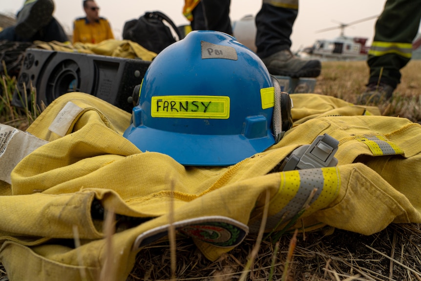 A blue helmet with "Farnsy" written on it sits on top of a yellow fire jacket in a field. Firefighters sit in the background.