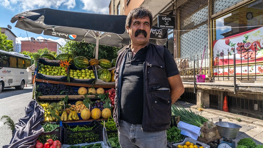A man with a thick black moustache stands with hands behind his back in front of a fruit stall with bananas, melons and apples
