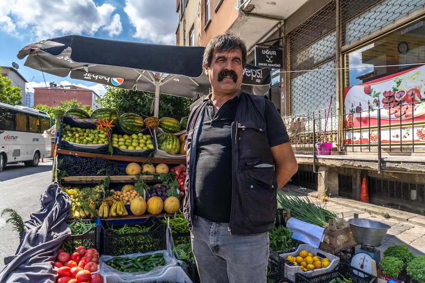 A man with a thick black moustache stands with hands behind his back in front of a fruit stall with bananas, melons and apples