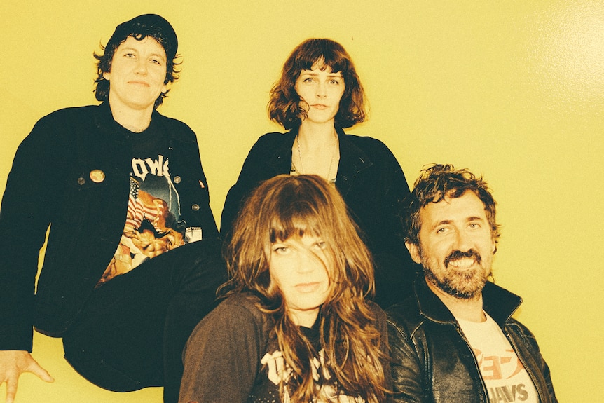 Three woman and one man, mostly wearing black, sit in front of a yellow background