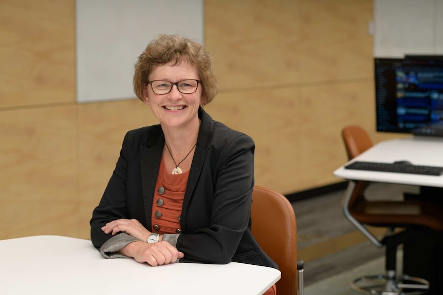 A woman with short brown hair wearing glasses and a black blazer sits at a table in a classroom.