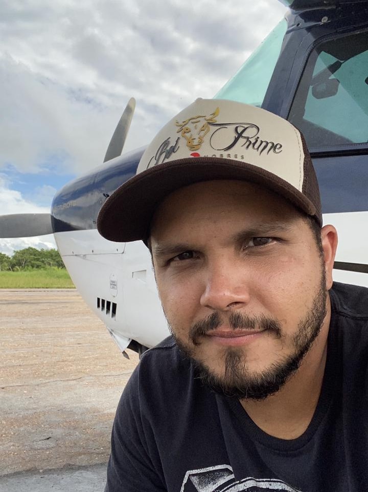  A man in a black shirt and baseball hat poses for a selfie in front of a plane 