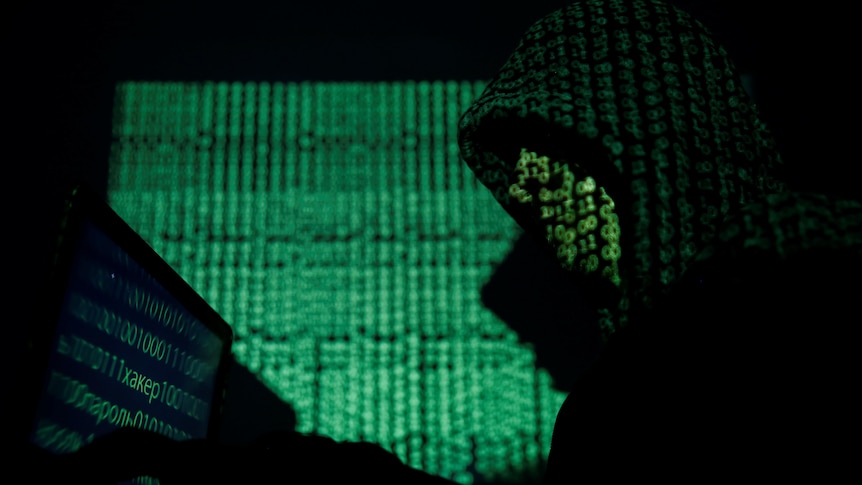 A hooded man holds a laptop computer as cyber code is projected on him.