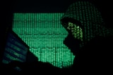 A hooded man holds a laptop computer as cyber code is projected on him.