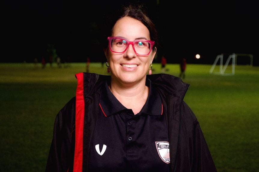 A woman with red glasses smiles with soccer fields behind her.