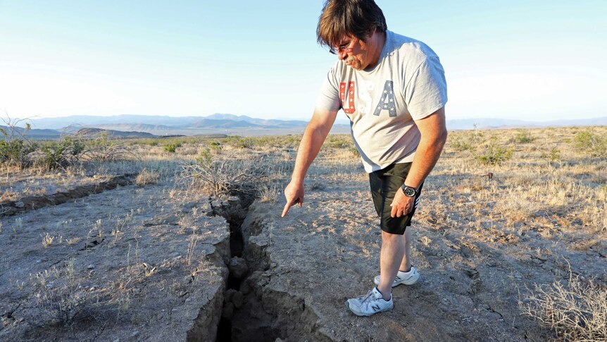 A man stands in a desert landscape and points to where a fissure has opened in the ground.
