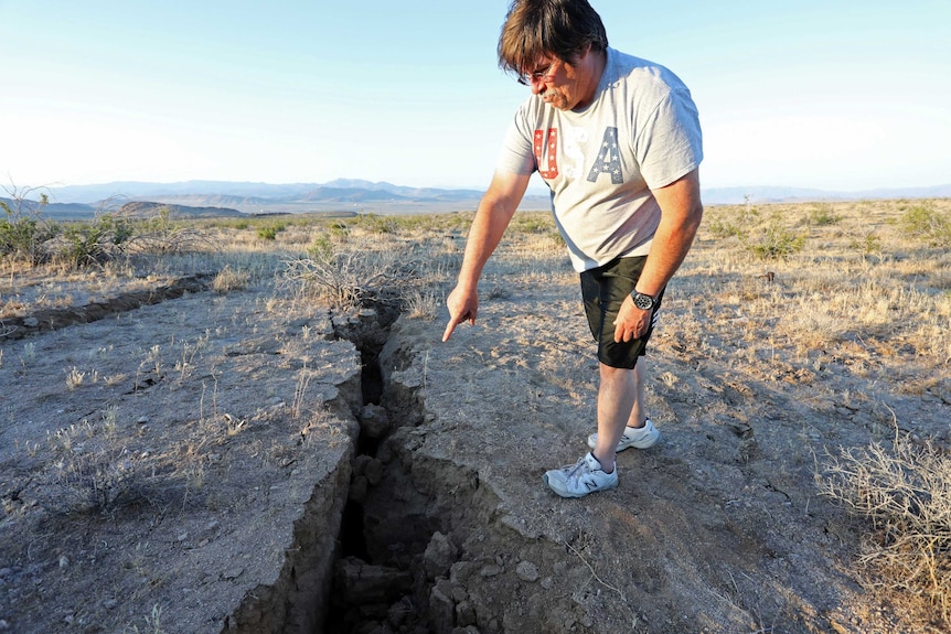 A man stands in a desert landscape and points to where a fissure has opened in the ground.