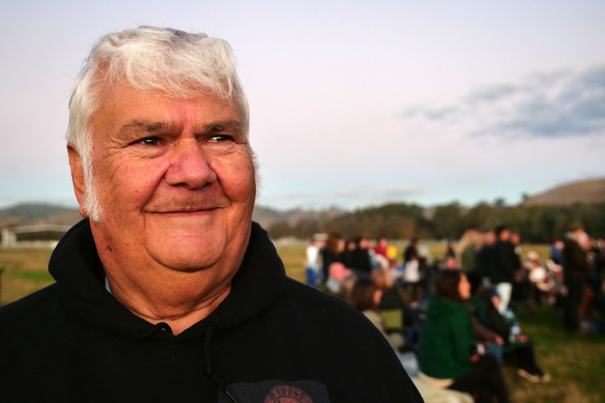 A man with short white hair smiles with a crowd behind him and rolling hills.