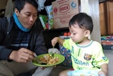 An Asian man sits on a small stool next to his young son, who has his spoon in his father's bowl of food.