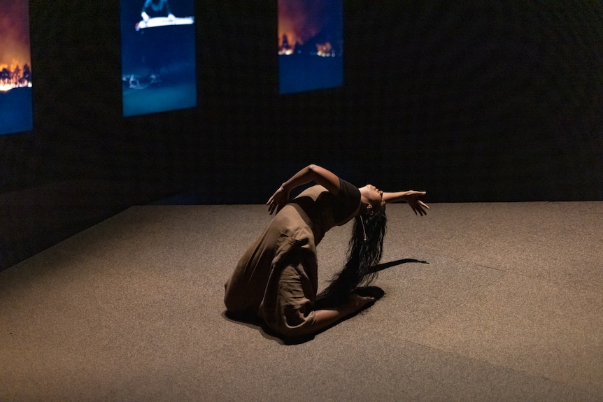 An Aboriginal woman on a slanted stage leans into a backbend, one arm stretched over her head