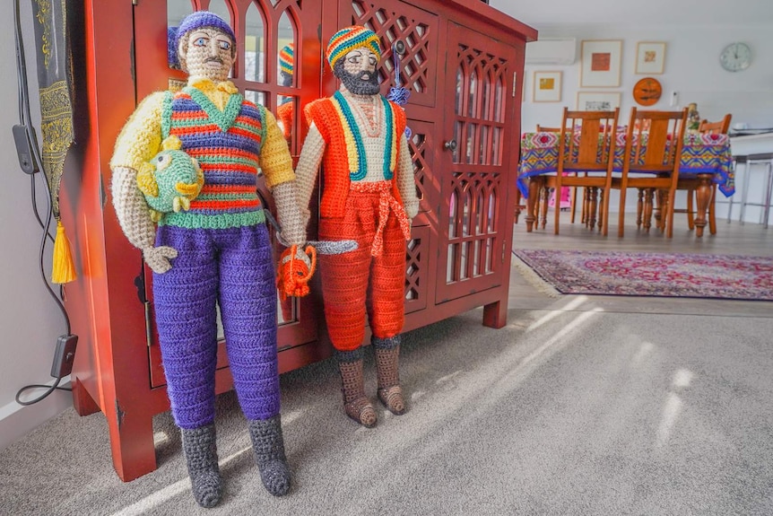 Two metre-tall crochet men in colourful costumes and headwear stand against a wooden dresser in a living room.