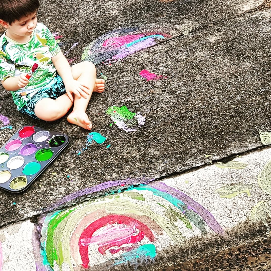 A small child sits on a concrete footpath, using homemade chalk to paint rainbows.