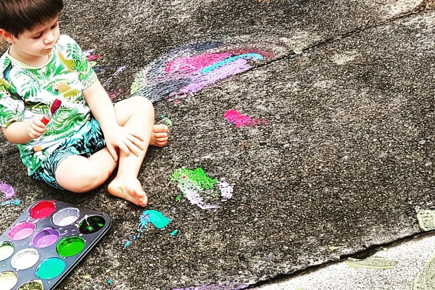 A small child sits on a concrete footpath, using homemade chalk to paint rainbows.