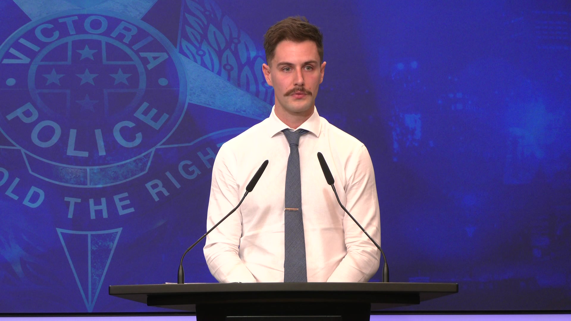 A man in a shirt and tie in front of a victoria police logo
