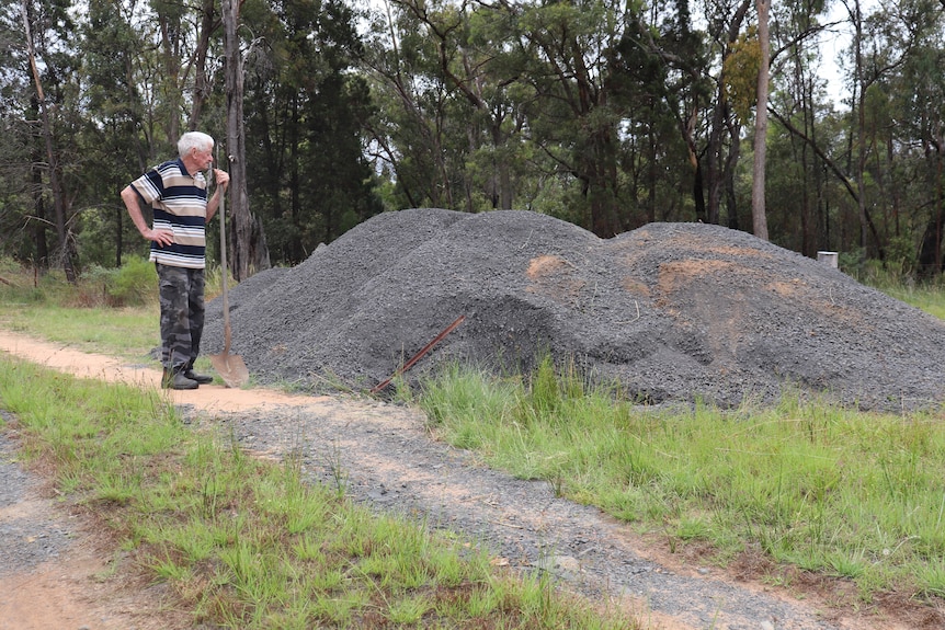 An elderly man stands with a shovel looking at a large pile of gravel at the entrance of his driveway