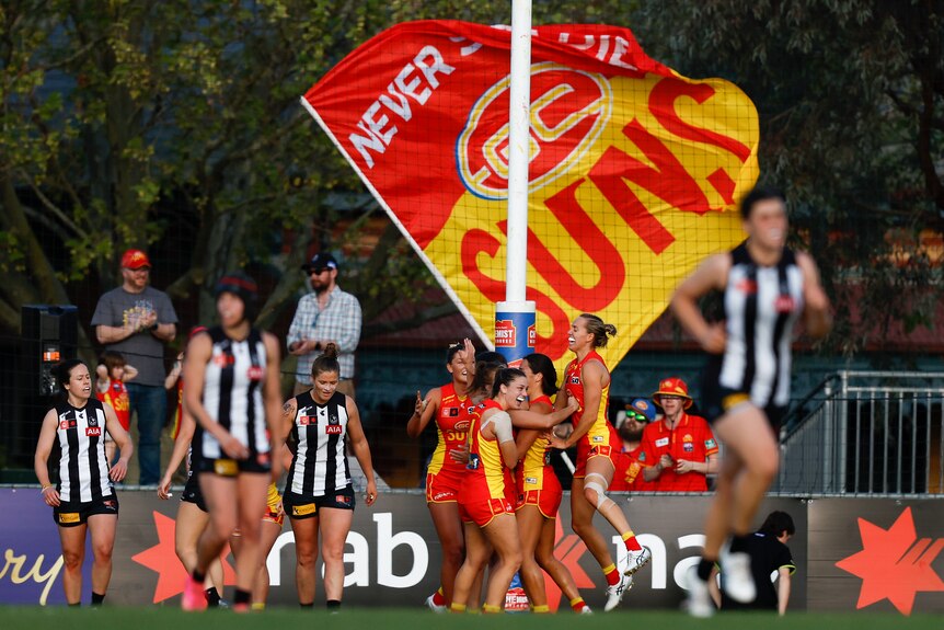 A group of Gold Coast Suns AFLW players embrace in the goalsquare after a goal, as fans wave a giant Suns flag behind the goals.