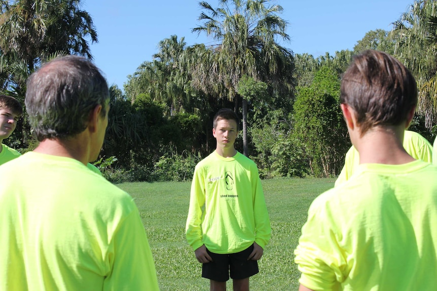 A teenage boy stands as part of a circle on the grass outdoors and watches on with his hands in his pockets.