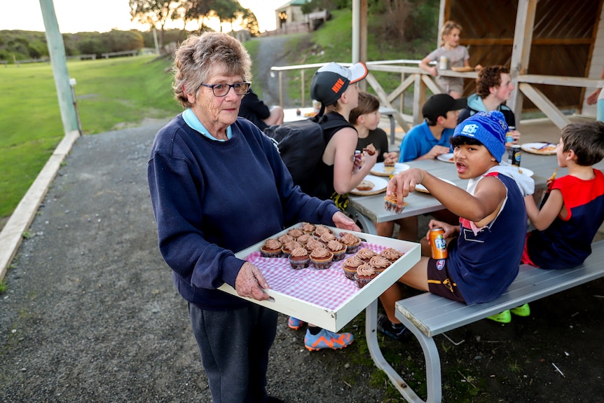 Older woman holding tray of cupcakes next to a football ground with kids seated next to her
